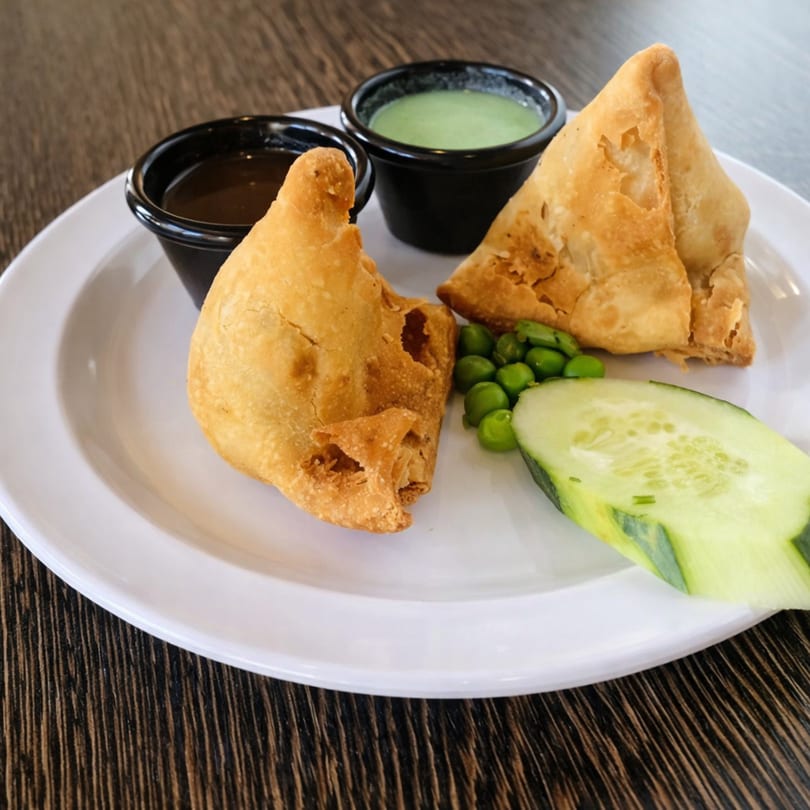 Two golden-brown Indian samosas with green peas, a cucumber slice, and two dipping sauces on a white plate.