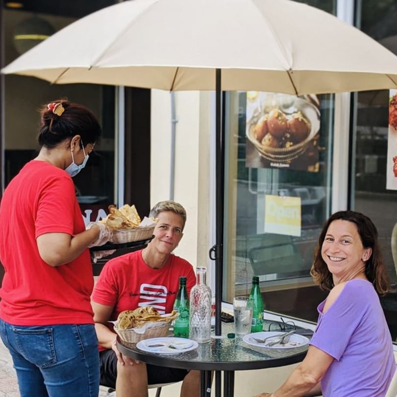 A server wearing a mask brings a basket of naan to two women dining outdoors at Sajna Indian Cuisine.