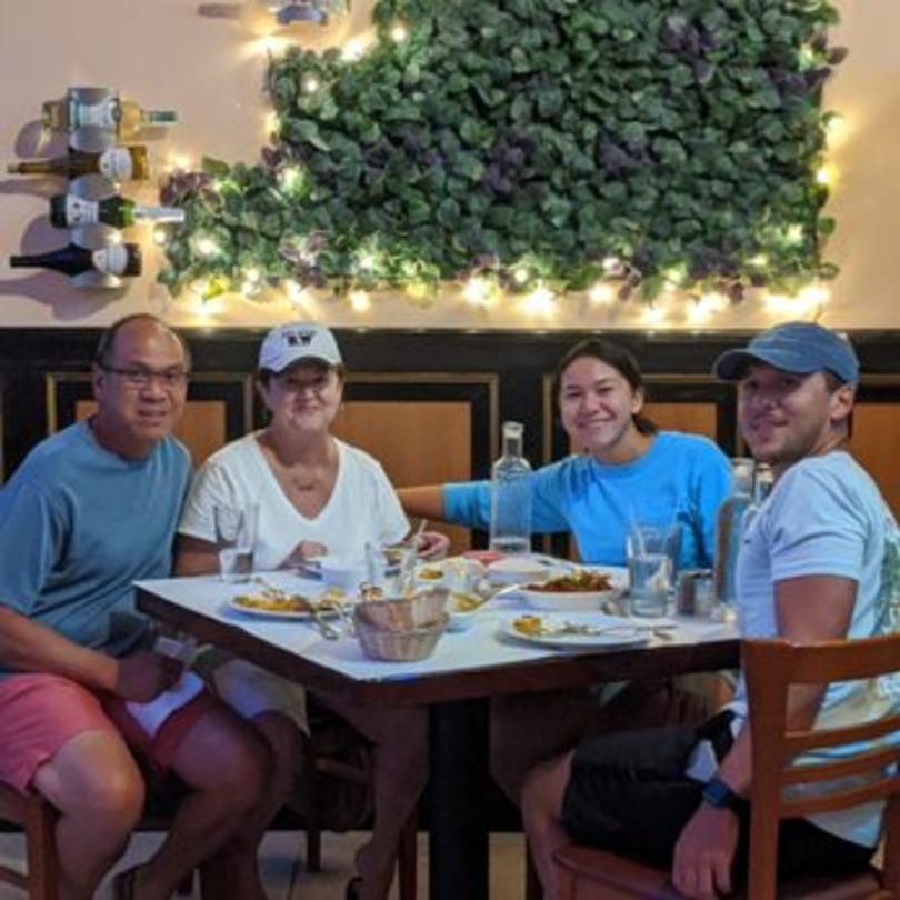 Four people smiling at a table with Indian food, wine bottles, and string lights in Sajna Indian Cuisine.