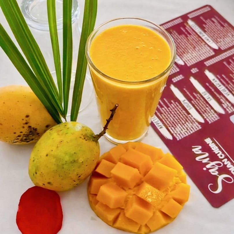 A glass of mango lassi with whole and diced mangoes, and a Sajna Indian Cuisine menu.