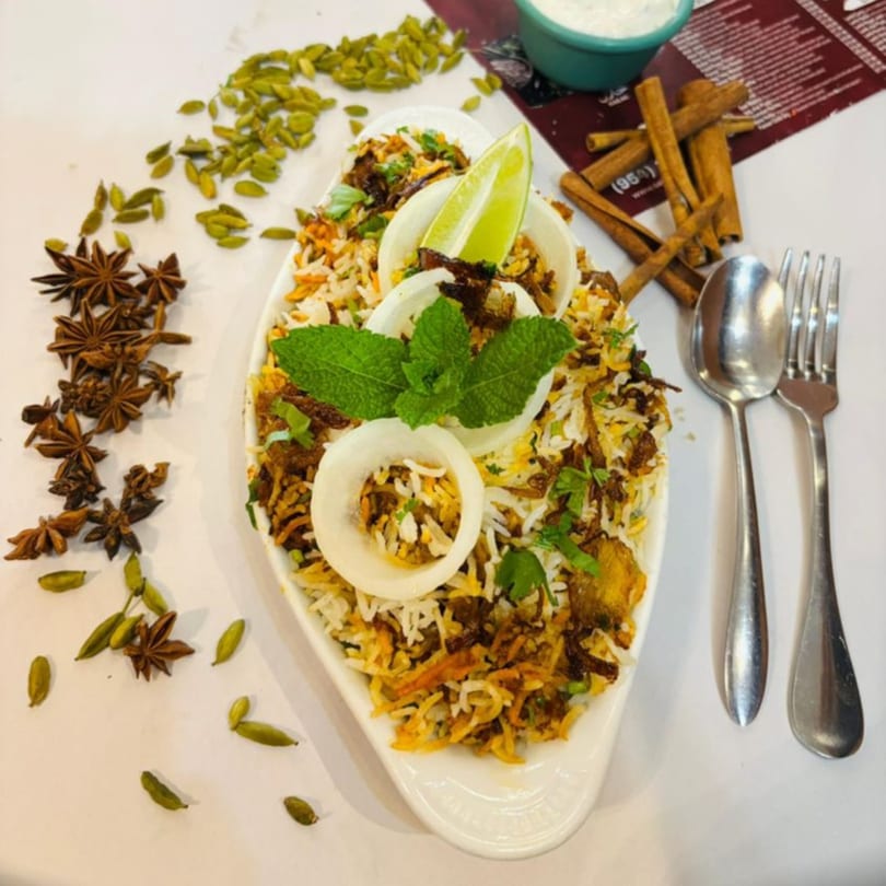 Sajna Indian Cuisine's biryani, garnished with mint, onion, and lime, surrounded by spices and cutlery.