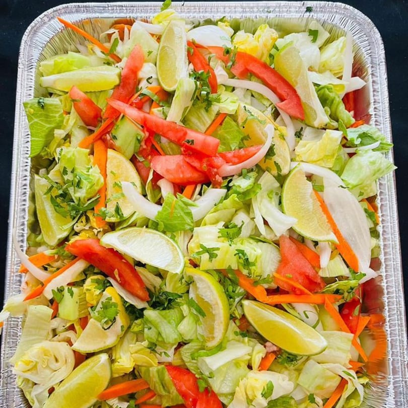 Fresh Indian salad with lettuce, tomatoes, carrots, onions, cilantro, and lime wedges in a foil tray.