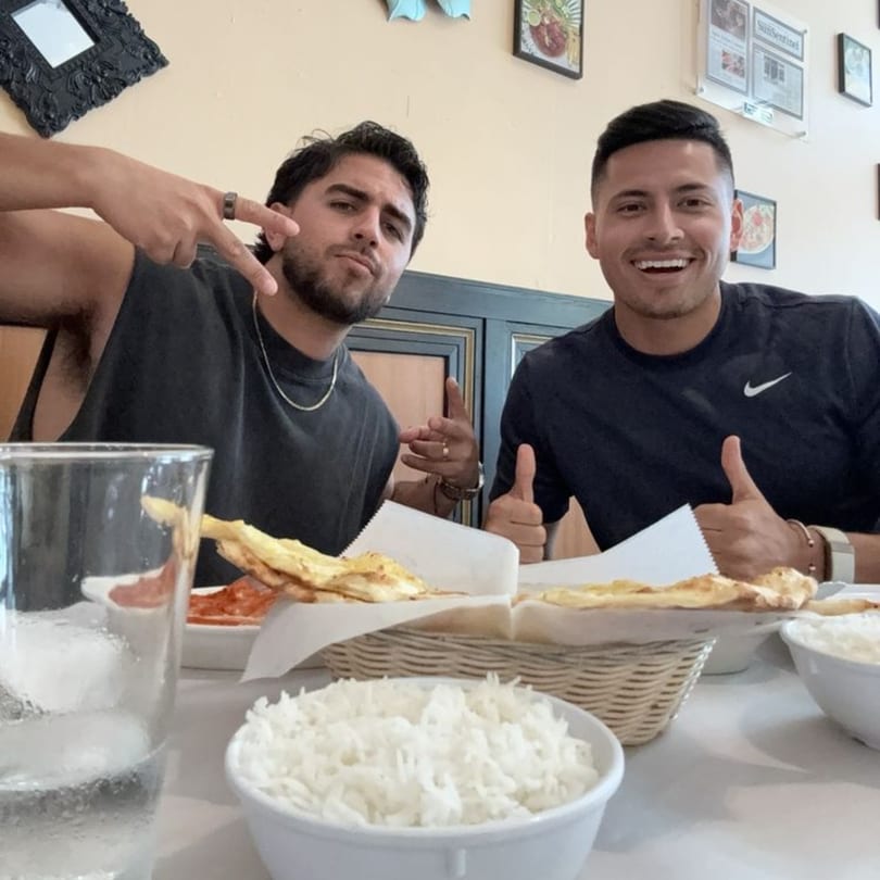 Two men smiling at Sajna Indian Cuisine, with bowls of rice and naan bread on the table.