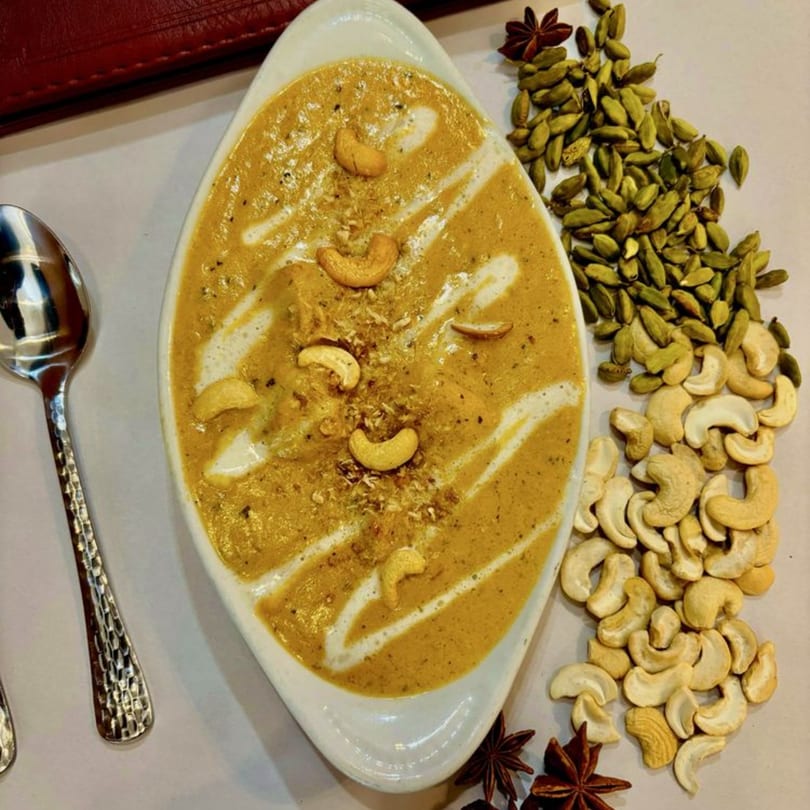 Sajna Indian Cuisine's creamy cashew curry, garnished with whole cashews and spices, next to a spoon.